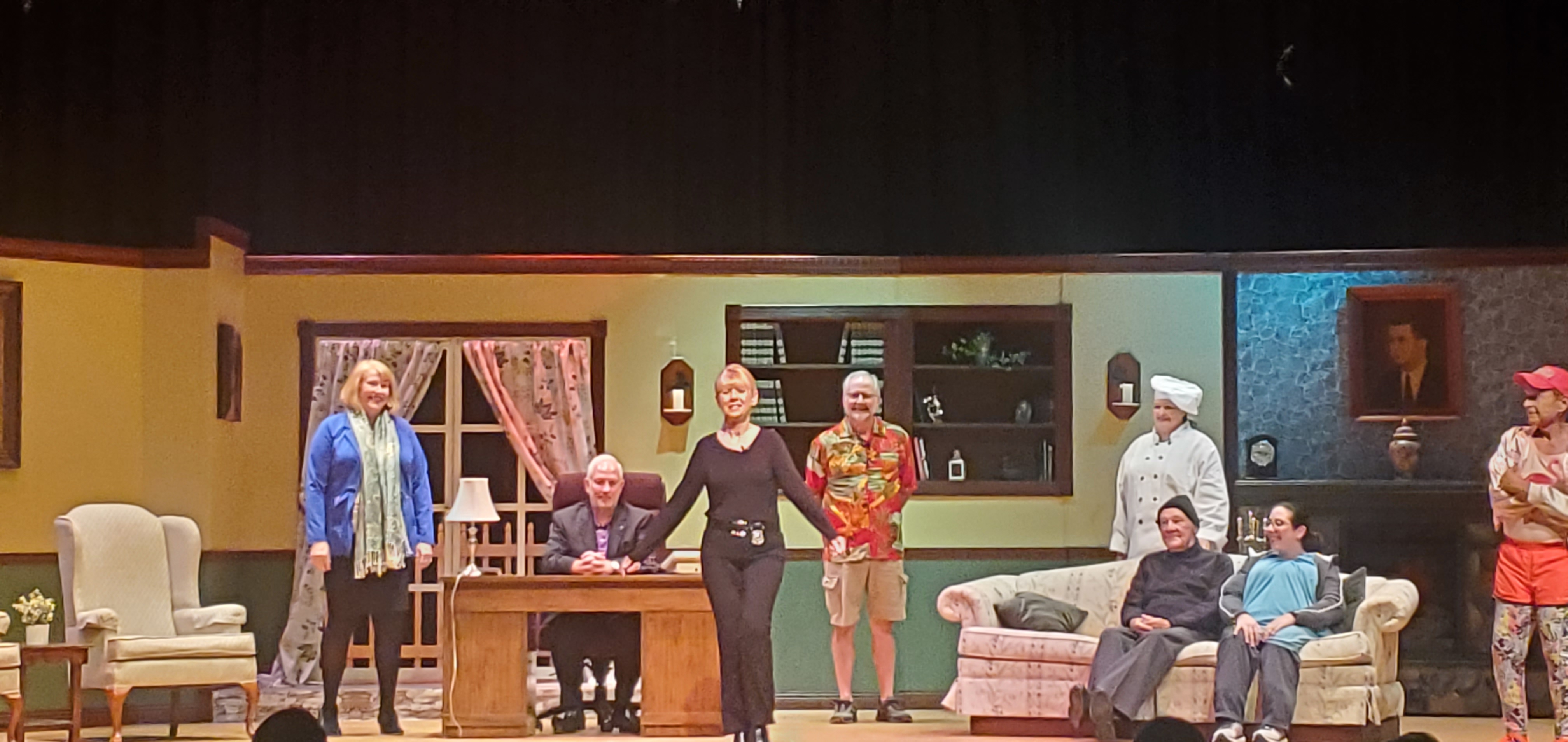From left to right: a woman in a blue jacket and light green scarf, a man in a black suit sitting behind a desk; a woman in a black top and pants standing in front of the desk; a man in a red Hawaiian shirt and shorts; a man in a black
       pullover, gray pants and black cap and a woman in a long bluish green top sitting on a couch; a chef standing behind the couch; and a man in an orange running outfit.