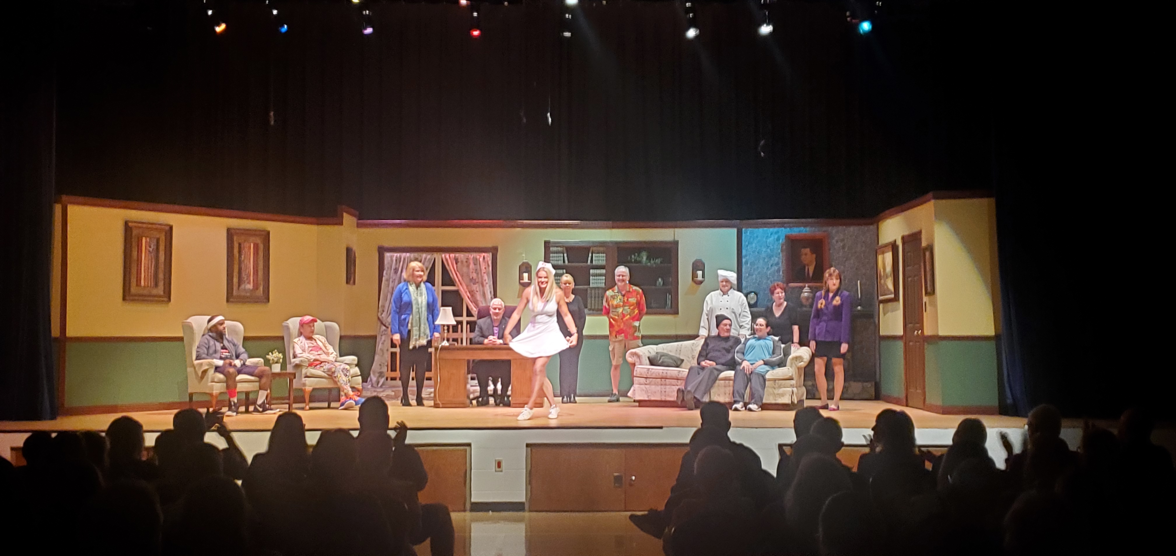 From left to right: a man wearing a white headband, purple shorts and a gray jacket sitting in a chair; a man in an orange running outfit sitting in a chair; a woman in a blue jacket and light green scarf standing; a man in a black suit sitting behind a desk, a woman
       wearing a short white skirt curtsying in front of the desk; a woman in a black top and pants standing to the right of the desk; a man in a red Hawaiian shirt and shorts; a man in a black pullover, gray pants and black cap and a woman in a long bluish green top sitting on a
       couch; a chef, a redheaded woman in a black top; and a woman in pigtails wearing a purple jacket and black skirt standing behind the couch.