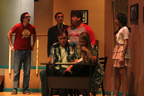A man and a woman sit at a desk, while a man in a red Avengers T-shirt, a man in a suit, a man in a Tampa Bay Buccaneers T-shirt
       and a young woman argue behind them
