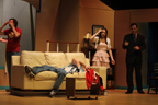 A man lies on the couch covering his face with his hands while a man in a red Avengers T-shirt, a young woman and
      man in a suit stand behind him