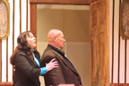 A women in a dark gray dress holds up a bald man with a head injury