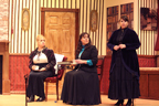 A woman in a white blouse and dark jacket and the woman in a dark dress and blue blouse sit while a woman in a long dark
       dress talks 