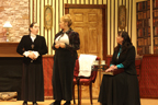 A woman in a black blouse talks to the woman in the white blouse and dark jacket while the woman in the dark dress and blue
       blouse sits and listens