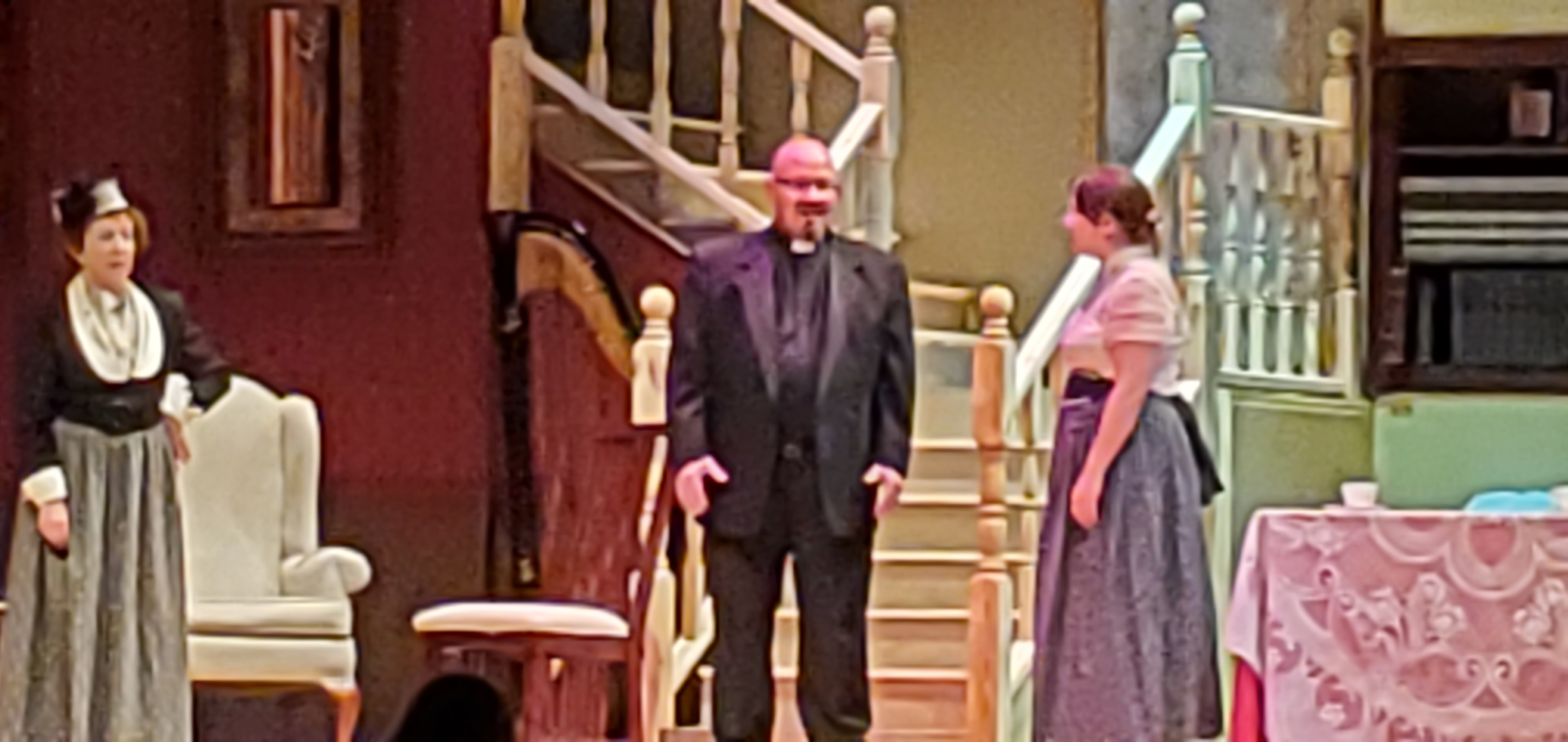 A woman in Victorian dress is to the left, the priest is in the middle and another woman in a white blouse and a long gray dress stands to the right.
       They stand in front of a staircase.