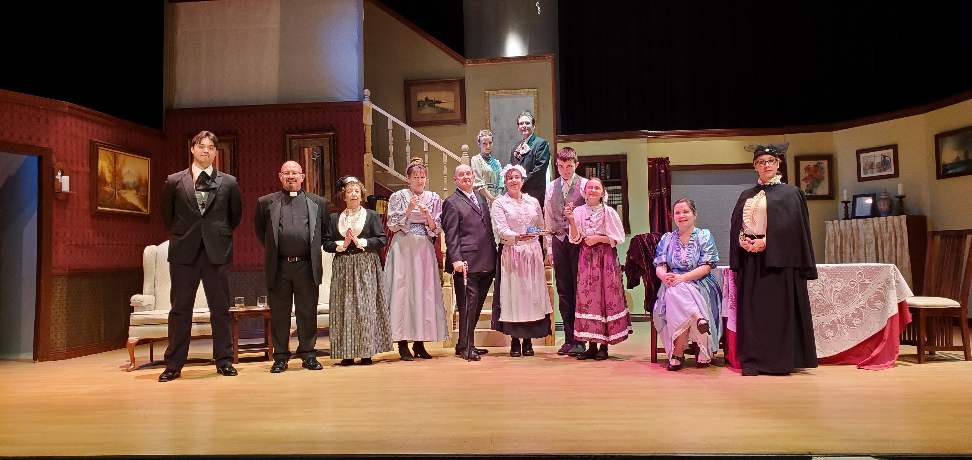 The entire cast standing, from left to right: a man in a black suit and ascot, a priest, a woman in Victorian dress, a woman in a long gray dress
       a man in a suit holding a cane, a maid in a white outfit, a tall boy in a white shirt and gray vest, a girl in a light rose top and rose colored dress, a woman in a metallic blue
       colored dress sitting, a woman in a black cloak and black dress standing. A teenage girl in a light green dress and a teenage boy in a gray suit stand behind and up on the
       landing.