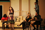 A man in a Bob marley shirt on the couch, a woman standing, another woman sitting, a man sitting reading a book, and another man stepping into the room from
       an open door